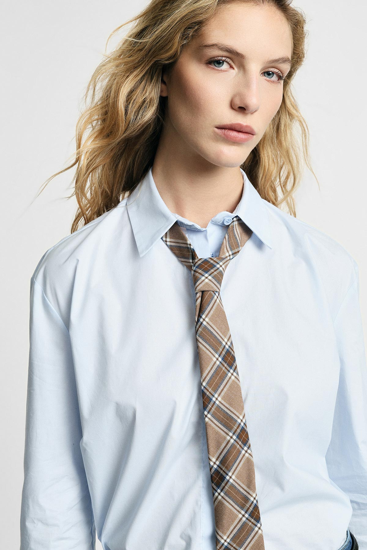 Blonde woman in light blue shirt and brown plaid tie, close-up fashion portrait with natural makeup and windblown hair