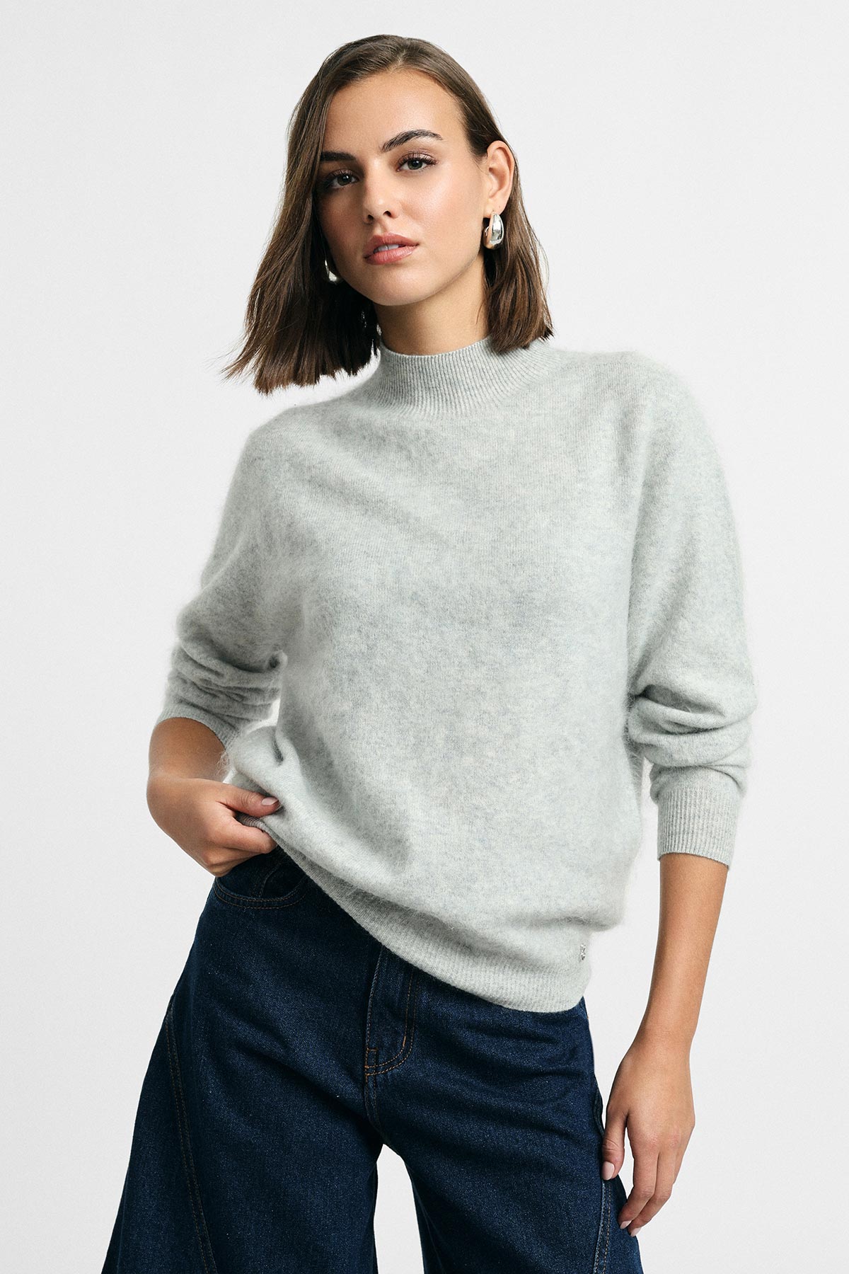 Woman modelling pale grey fuzzy sweater and dark blue jeans, standing with hand in pocket against a plain light background