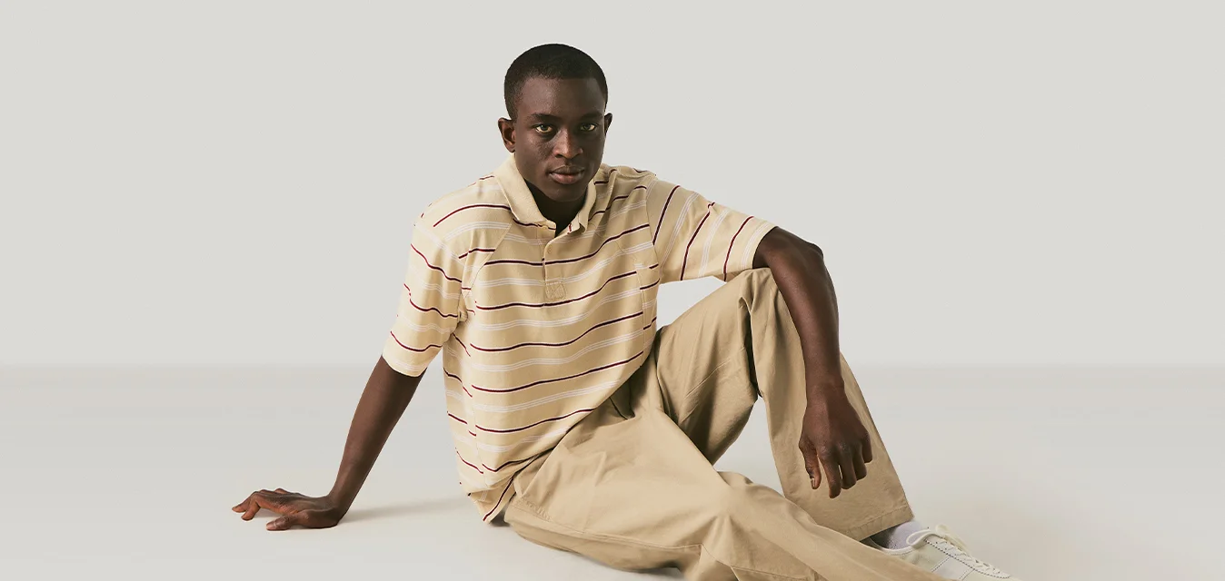 Giovane uomo seduto sul pavimento con un braccio disteso all'indietro, indossa una maglietta polo a righe, pantaloni beige e sneaker bianche.