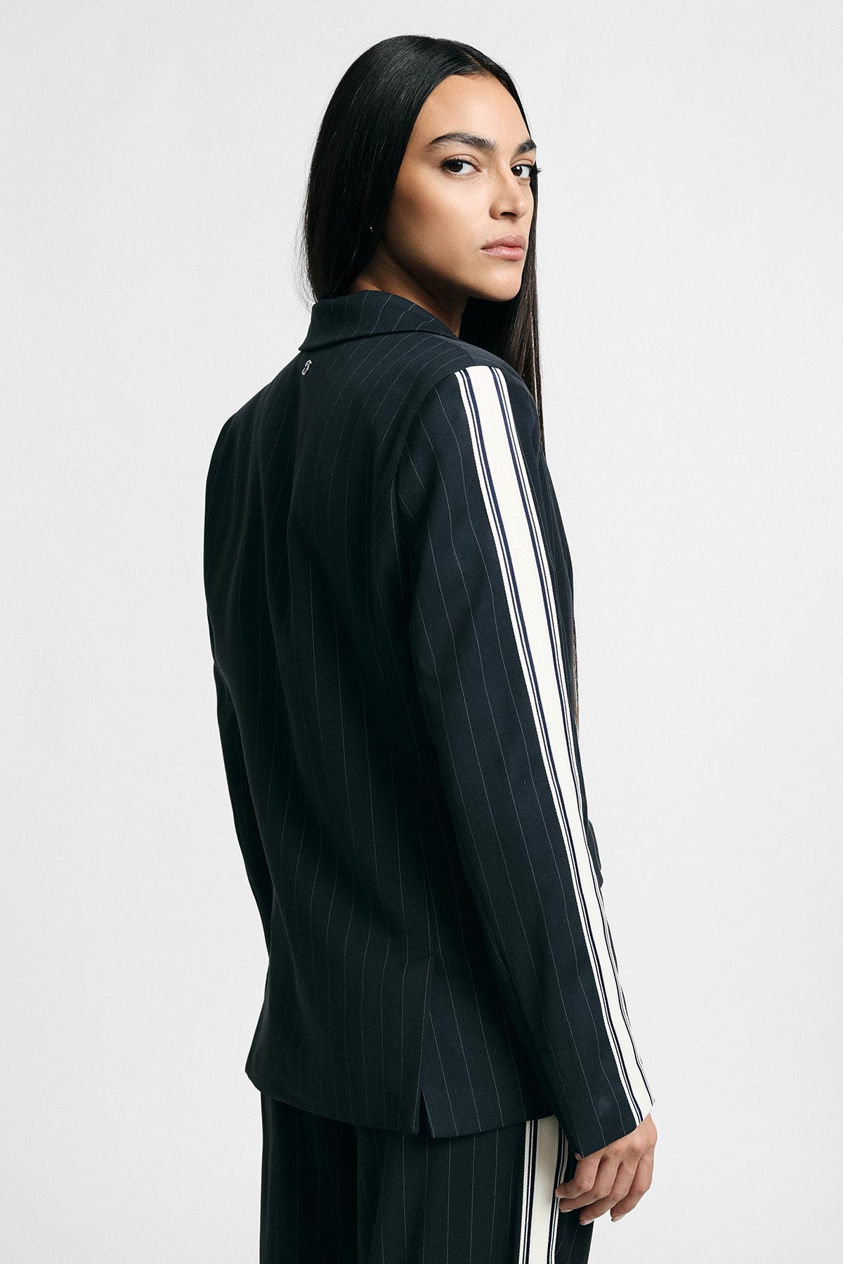 Woman with long dark hair wearing a navy pinstripe blazer with white side stripes, looking over her shoulder against a plain white background