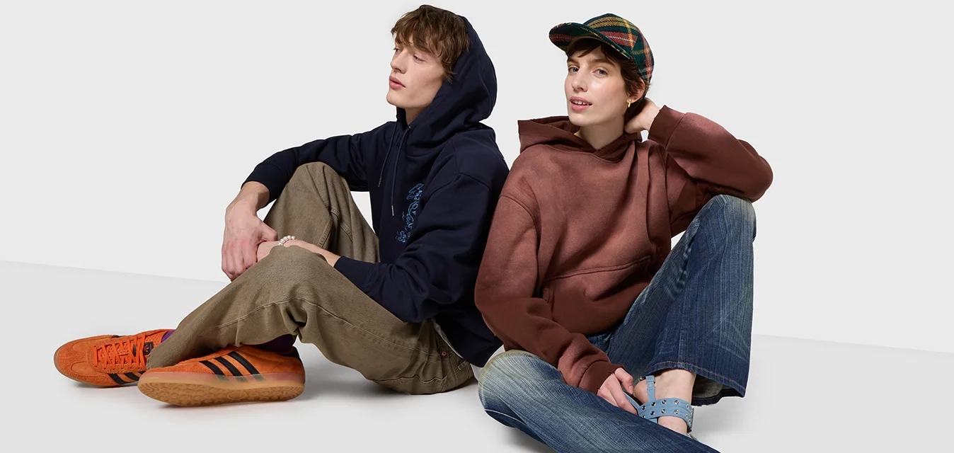 Due persone siedono su una superficie bianca, indossando un felpa blu marina, un felpa marrone, pantaloni khaki e jeans di denim, con sneaker arancioni e scarpe blu.