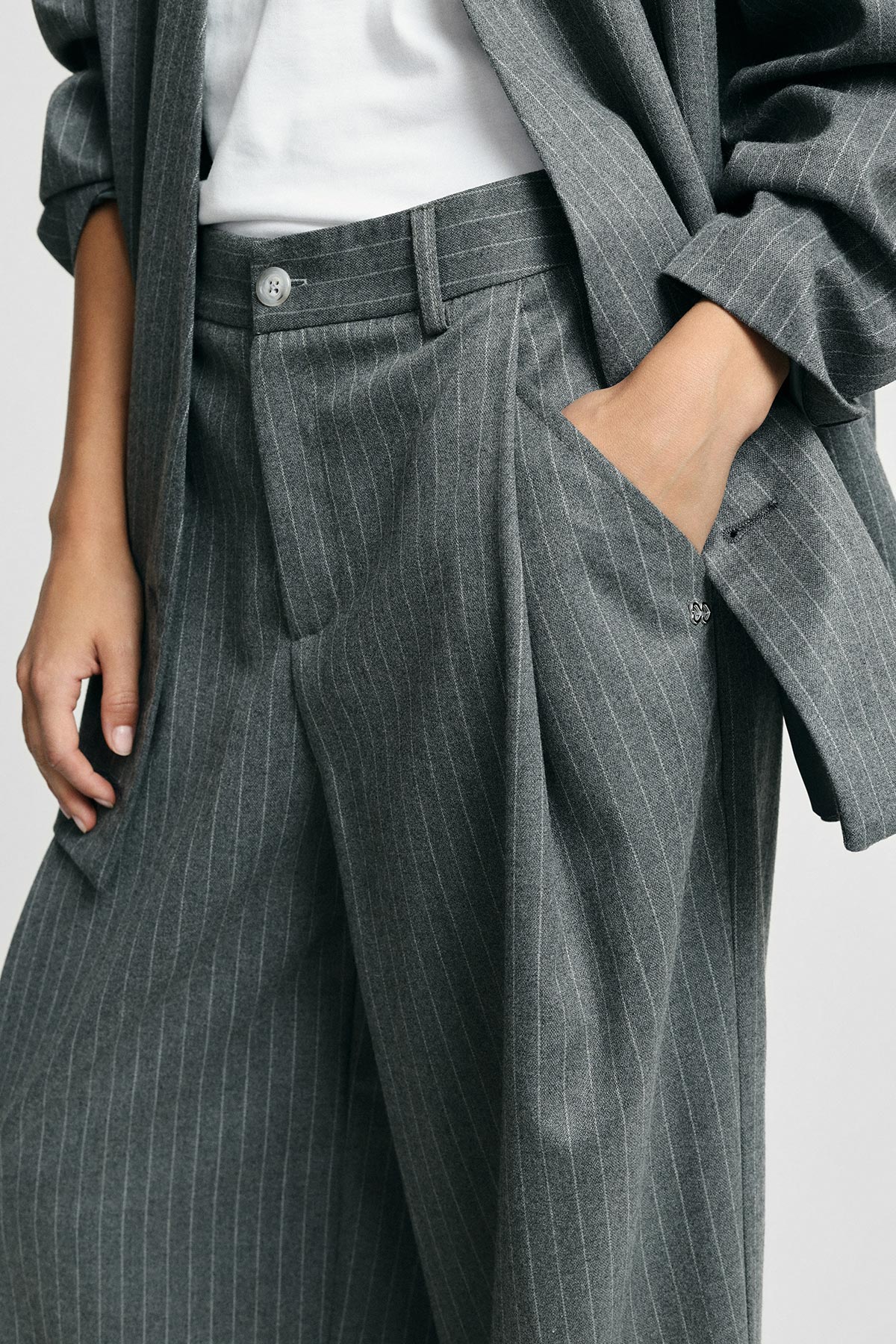 Close-up of person wearing grey pinstripe suit: high-waist pleated trousers and matching blazer, hand in pocket with white top visible