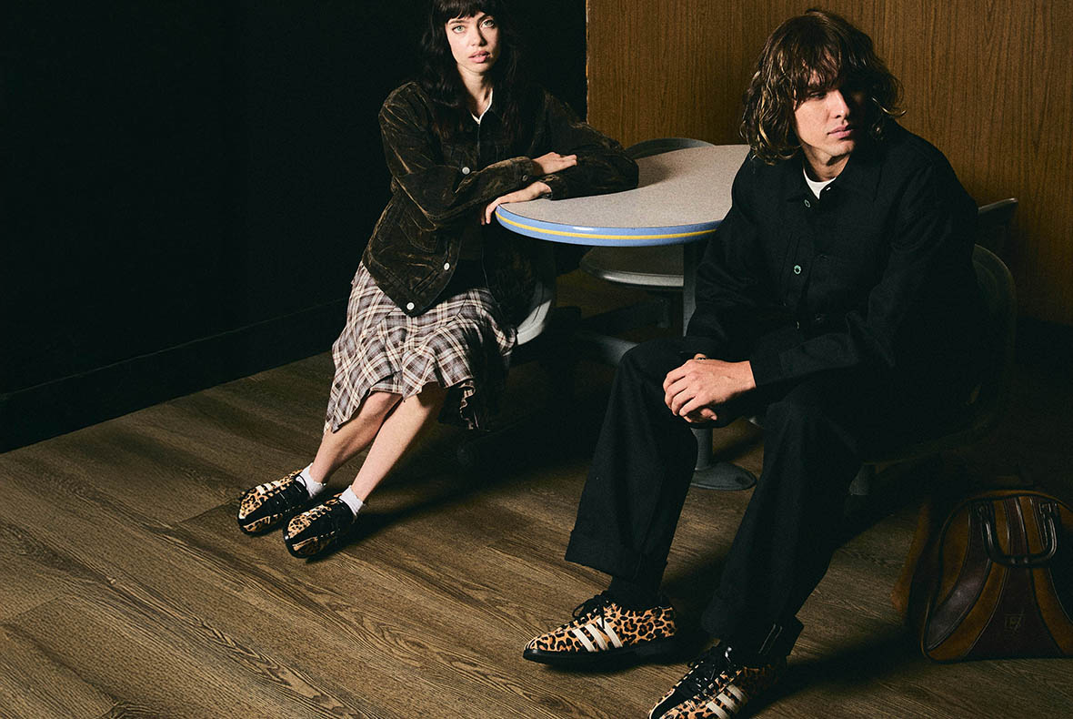 Two young adults seated at a retro round table on a wooden floor, wearing leopard-print trainers and vintage-inspired casual outfits