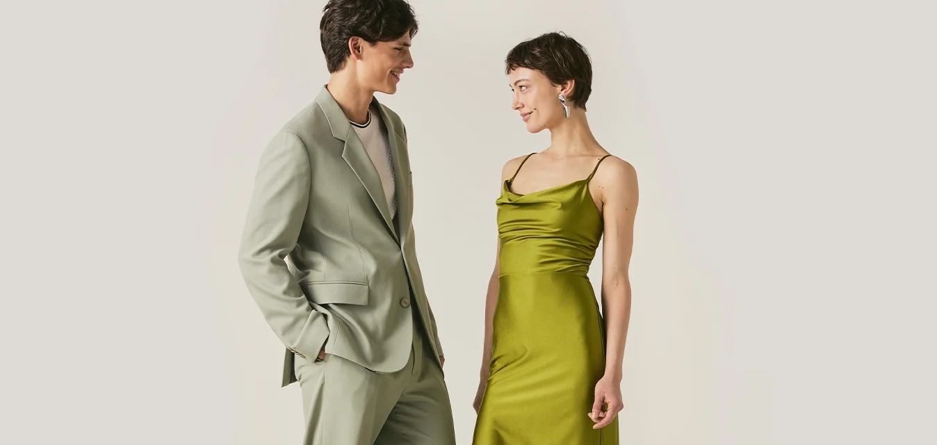 Uomo in abito verde chiaro e donna in vestito di raso verde con orlo in pizzo stanno uno di fronte all'altro, sorridendo, su sfondo semplice.