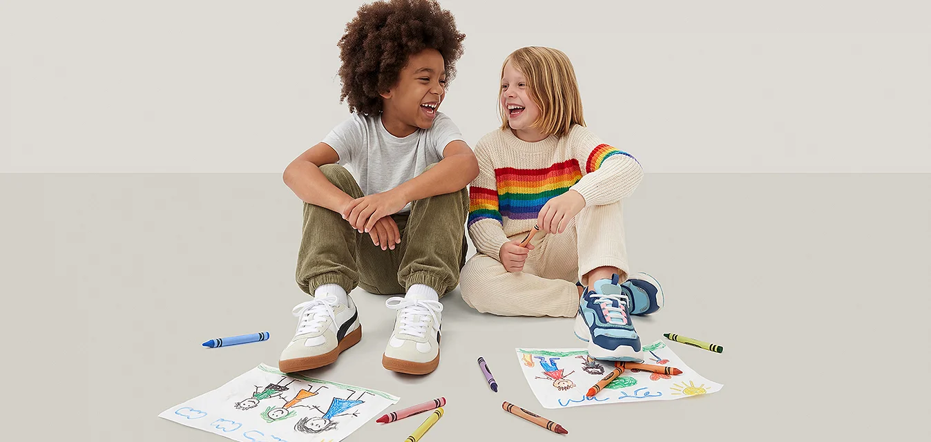 Due bambini seduti per terra, sorridenti e ridendo, con pastelli e disegni colorati sparsi davanti a loro.