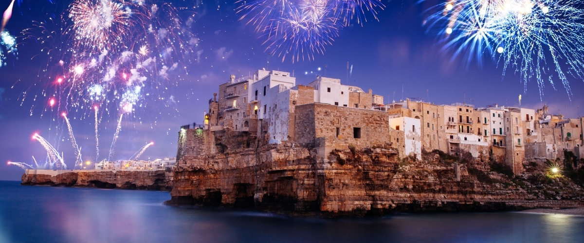 Fireworks over a cliffside Mediterranean coastal town at night, historic stone buildings lit and reflected in the calm sea