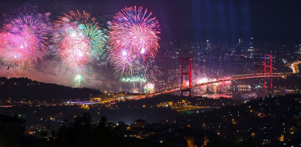 Night fireworks display over a lit suspension bridge spanning a river, colourful bursts and beams above the city skyline