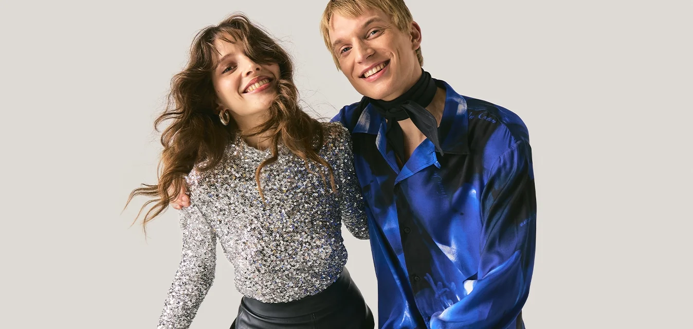 Giovane uomo sorridente in camicia blu e giovane donna in top scintillante e gonna nera posano insieme contro uno sfondo neutro.