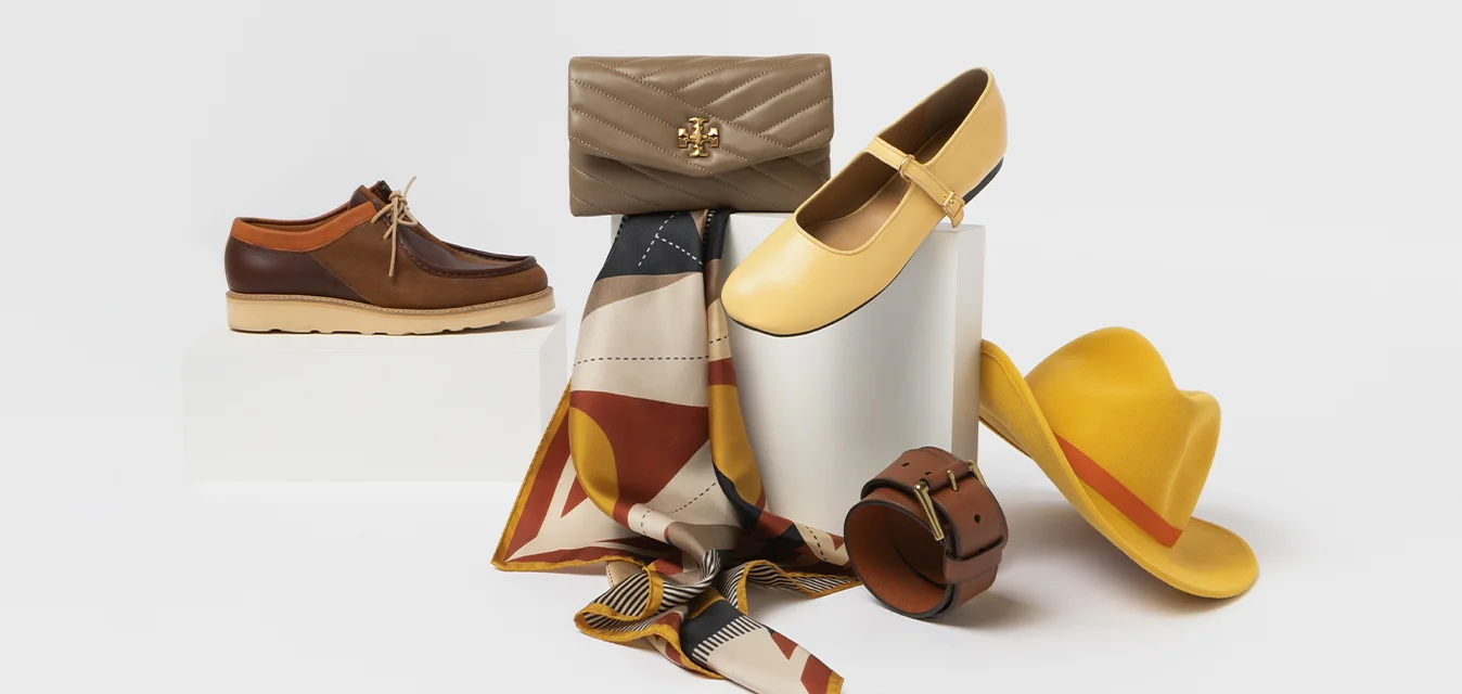 Scarpe marroni con tomaia in pelle, scarpe gialle basse, una borsa beige, una sciarpa a motivi, un cappello giallo e una cintura marrone, tutti esposti su uno sfondo bianco.