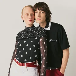 Due giovani adulti si tengono vicini; uno indossa una maglia trasparente a pois con pantaloncini bianchi, l'altro una polo nera e pantaloni rossi su uno sfondo semplice.