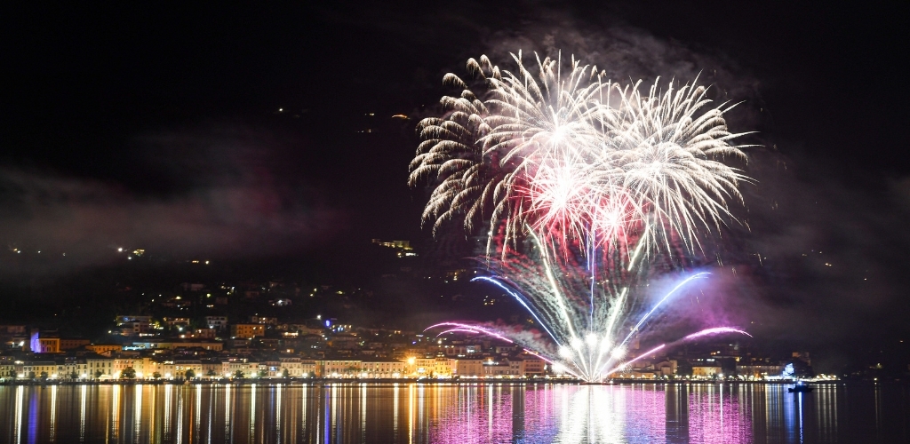 Nighttime fireworks display over a lit waterfront town, colourful bursts reflected across the calm harbour water