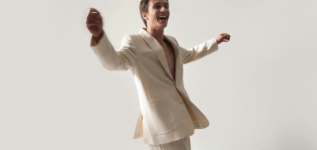 Uomo in completo beige chiaro sorride e alza le braccia mentre cammina davanti a uno sfondo grigio chiaro semplice.