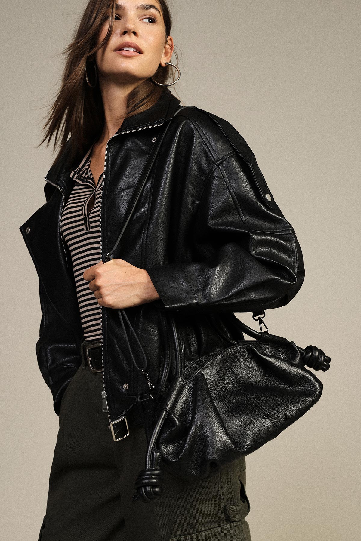 Model wearing oversized black leather biker jacket and striped top, holding slouchy black leather bag