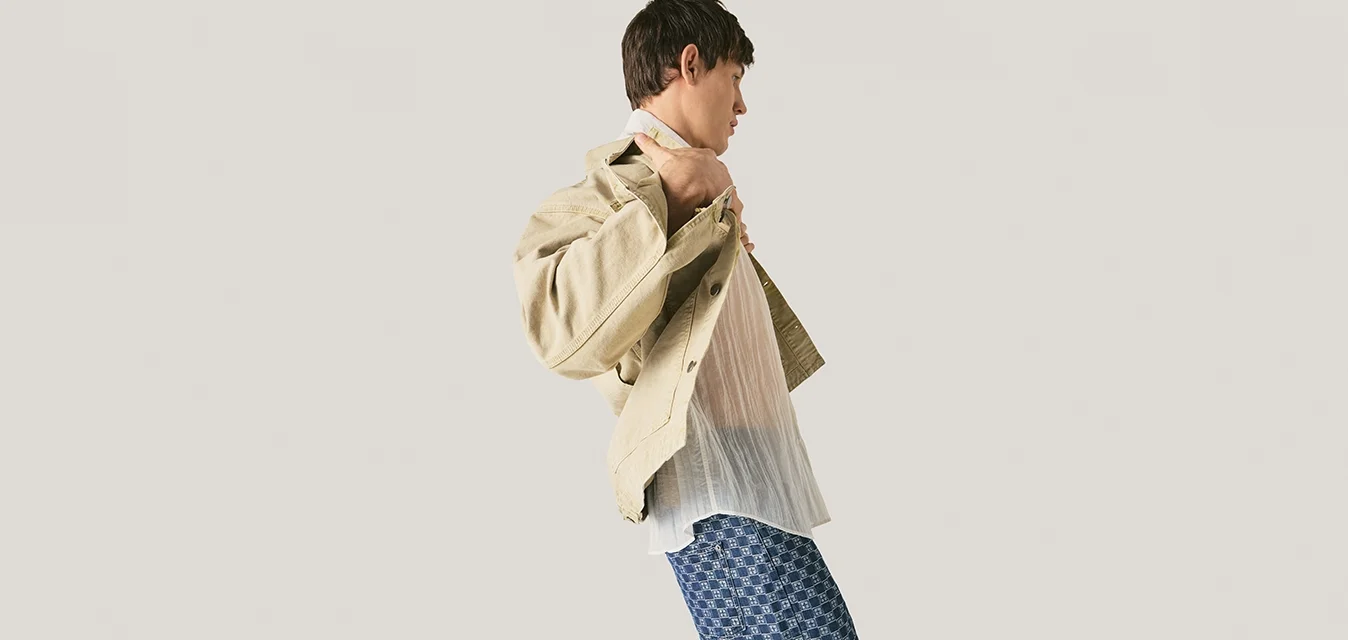 Giovane uomo che sistema una giacca beige sopra una camicia bianca, indossa pantaloni blu con motivi, in piedi contro uno sfondo grigio chiaro uniforme.