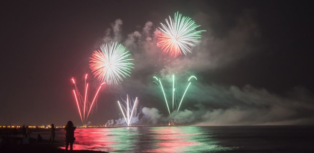 Red and green fireworks bursting over a calm sea at night with reflections on the water and silhouetted spectators on the beach