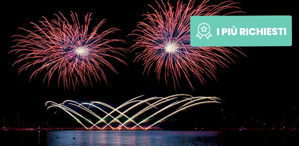 Night fireworks display over calm harbour with colourful bursts and arcing fountains reflected on water; teal badge reading 'I PIÙ RICHIESTI'.