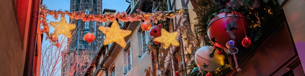 Narrow European street decorated with illuminated star garlands and oversized Christmas baubles, cathedral spire in background