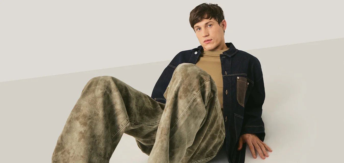 Giovane uomo seduto a terra con le gambe piegate, indossa pantaloni verdi larghi, camicia beige e giacca scura su uno sfondo neutro.