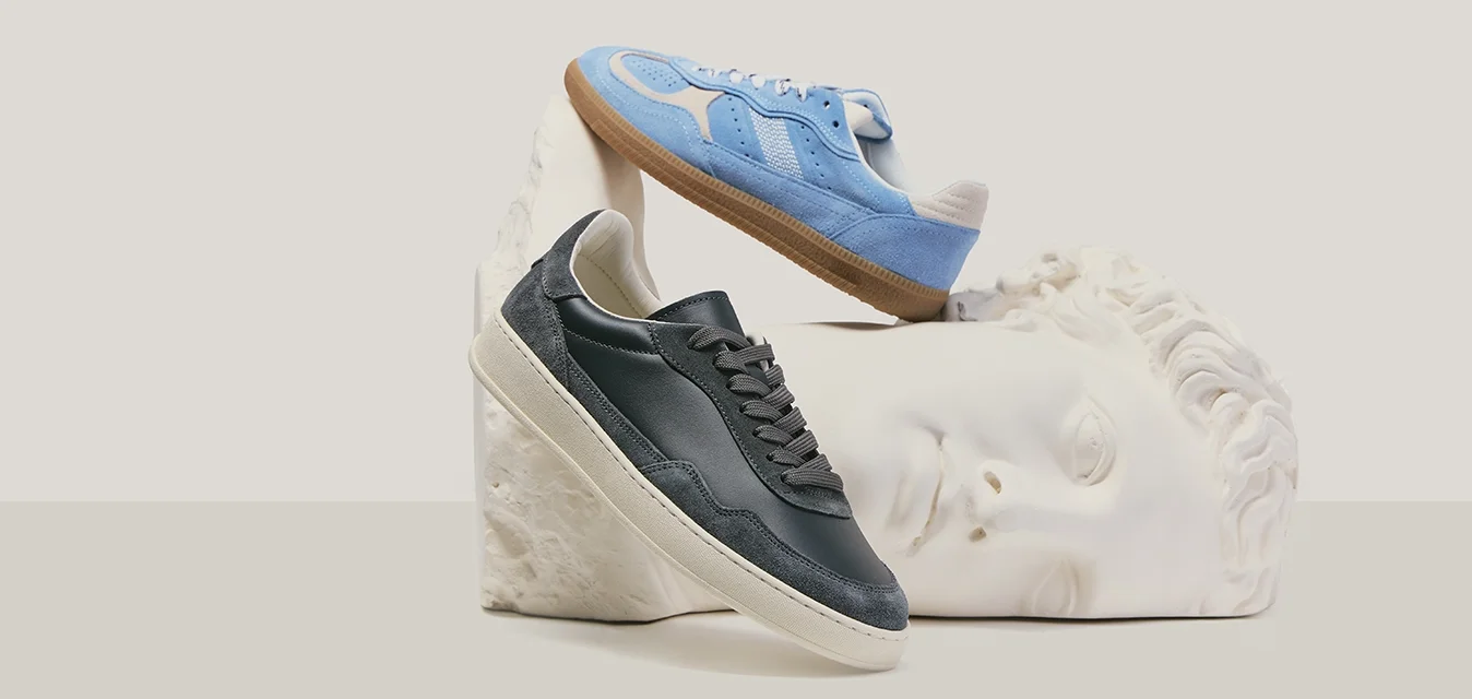 Due sneakers, una nera e una blu, esposte su e sopra una testa di scultura classica bianca contro uno sfondo neutro.
