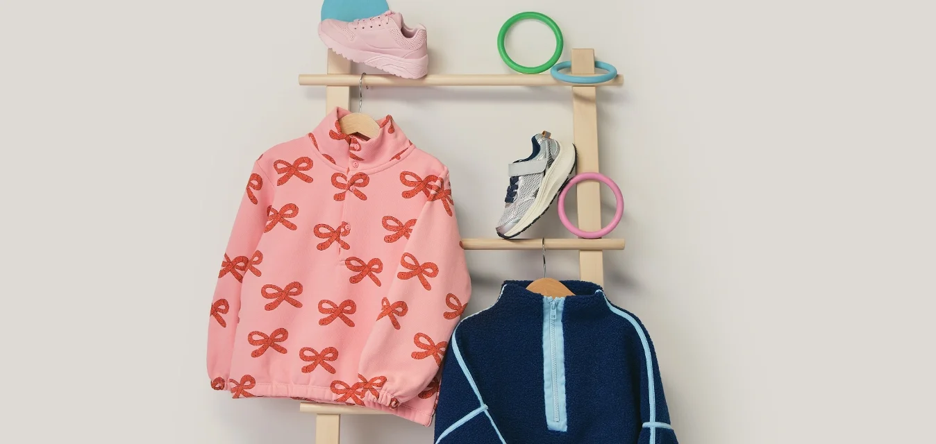 Due giacche per bambini, una rosa con fiocchi arancioni e l'altra blu navy con strisce azzurre, appese a un appendiabiti di legno con due paia di sneakers sopra.