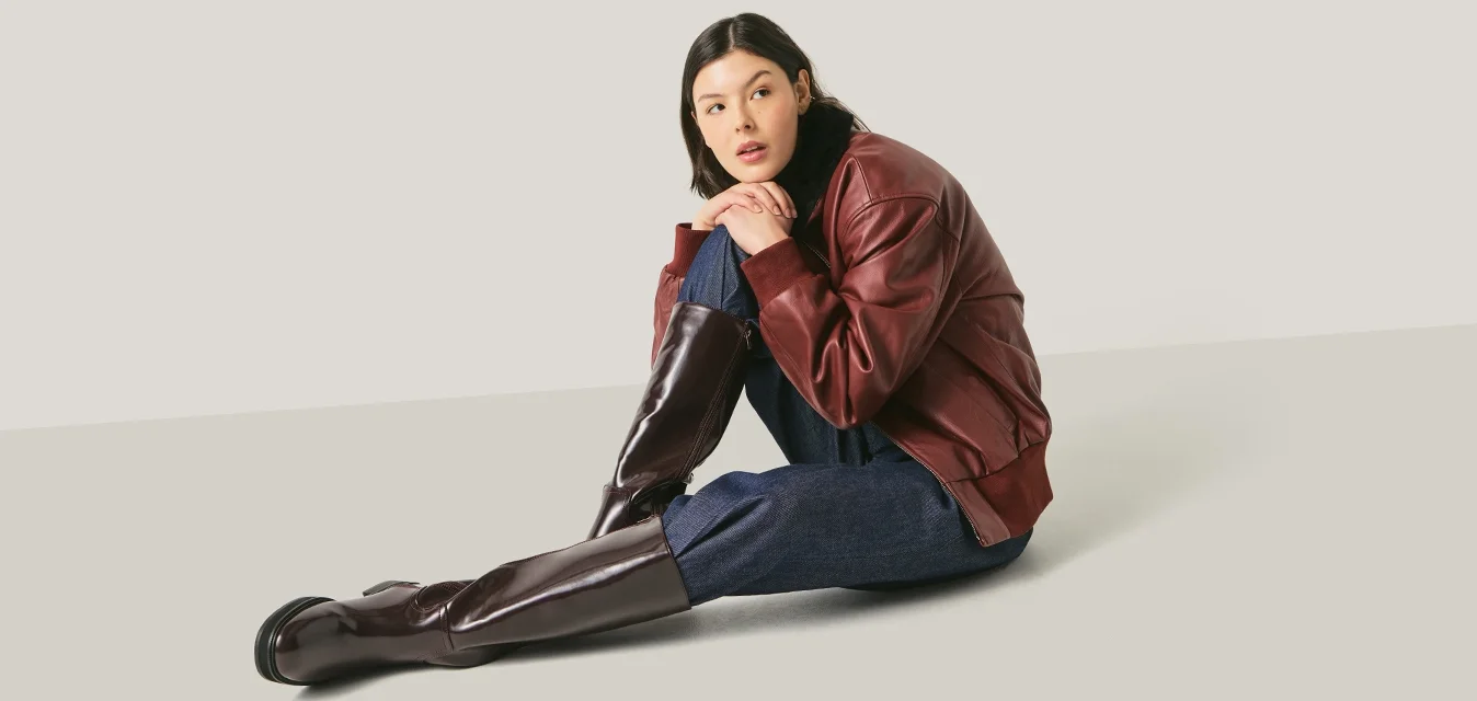 Donna con giacca di pelle rossa, pantaloni blu navy e stivali marroni alti seduta su una superficie chiara, inclinata in avanti con le mani intrecciate intorno a un ginocchio.
