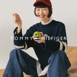 Giovane con un cappellino rosso, maglione blu navy e jeans seduto su un divano in pelle color cuoio, che tiene una mela verde sorridendo davanti a uno sfondo neutro.