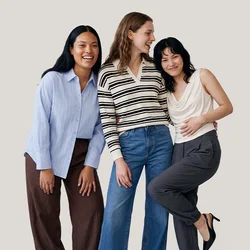 Tre donne in piedi molto vicine, sorridenti e che ridono, vestite con abiti casual su uno sfondo chiaro e uniforme.