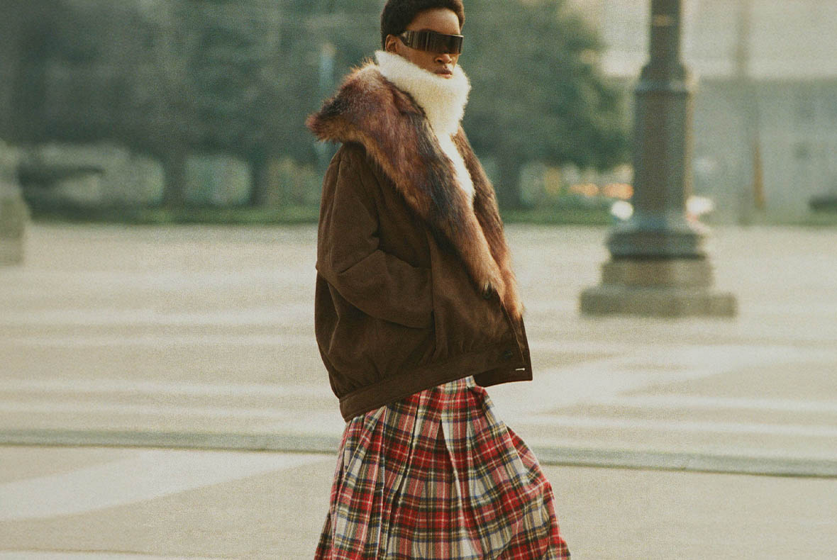 Person walking in city wearing oversized brown shearling jacket with fur collar, white scarf, tartan skirt and dark sunglasses
