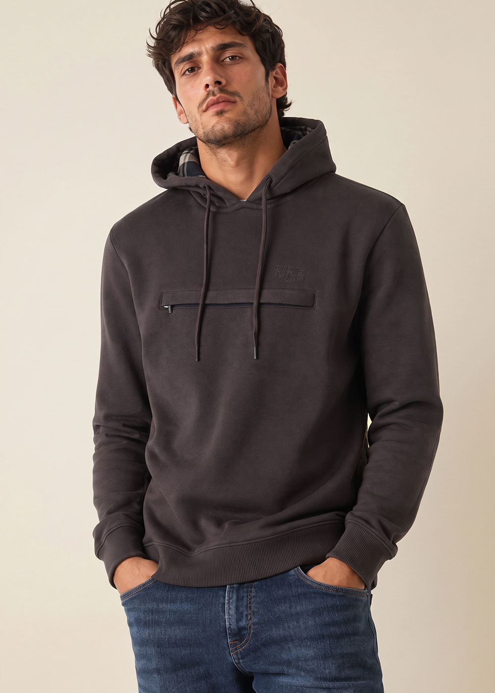 Male model wearing charcoal men's hoodie with chest zip pocket and drawstrings, hands in jeans pockets, neutral background