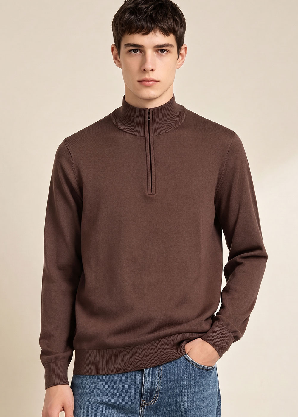 Young man modelling men's brown half-zip jumper and blue jeans, hand in pocket against neutral background