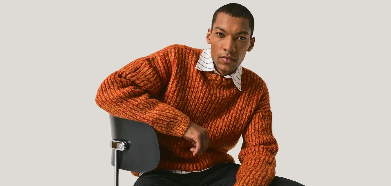 Uomo con maglione arancione e pantaloni neri seduto al contrario su una sedia nera contro uno sfondo semplice grigio chiaro.