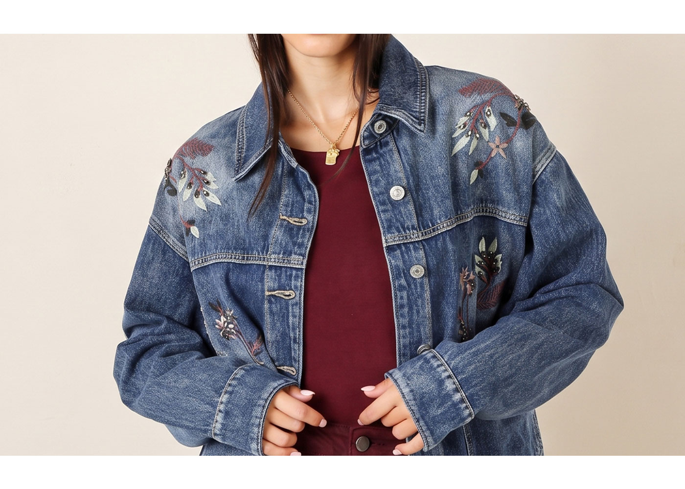Cropped view of embroidered blue denim jacket worn over a burgundy top with a small gold pendant necklace