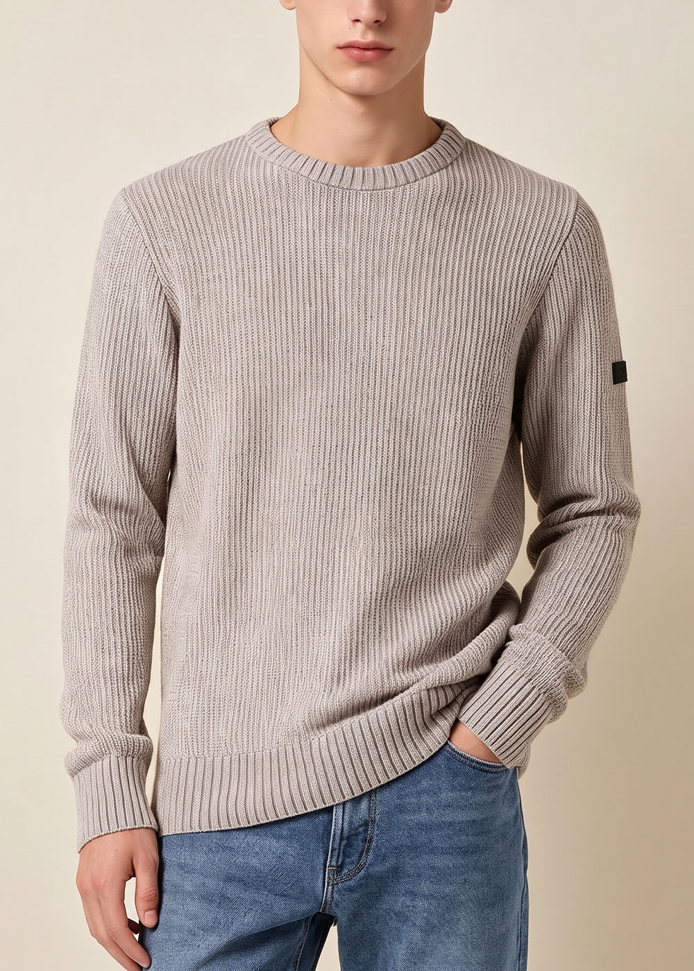 Male model wearing beige ribbed crew-neck sweater and blue jeans, casual pose with hand in pocket against a neutral background