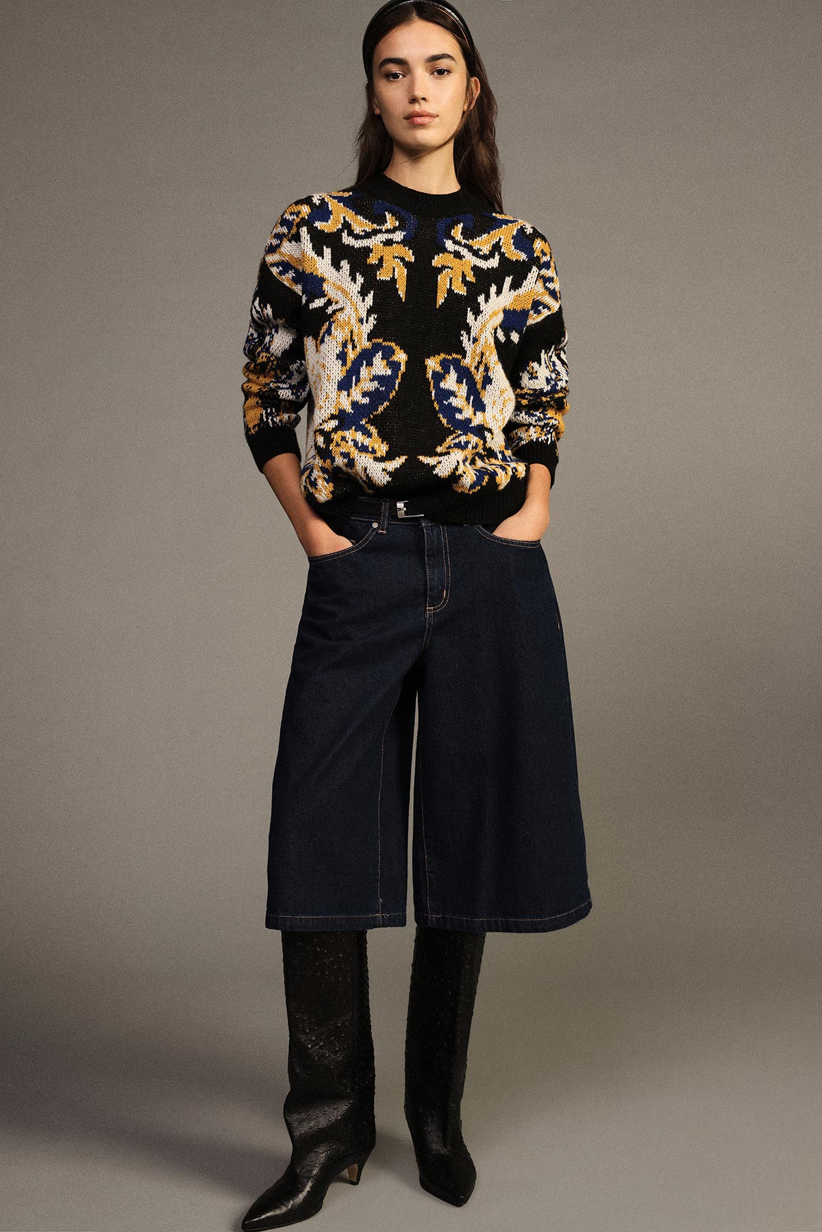 Young woman modelling patterned navy and gold knit sweater with wide denim culotte skirt and knee-high black boots, hands in pockets