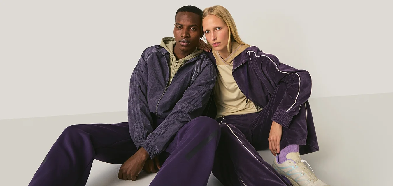 Due modelle sedute sul pavimento indossano tute viola coordinate, una con felpa beige con cappuccio e l'altra con maglietta beige e sneakers bianche.