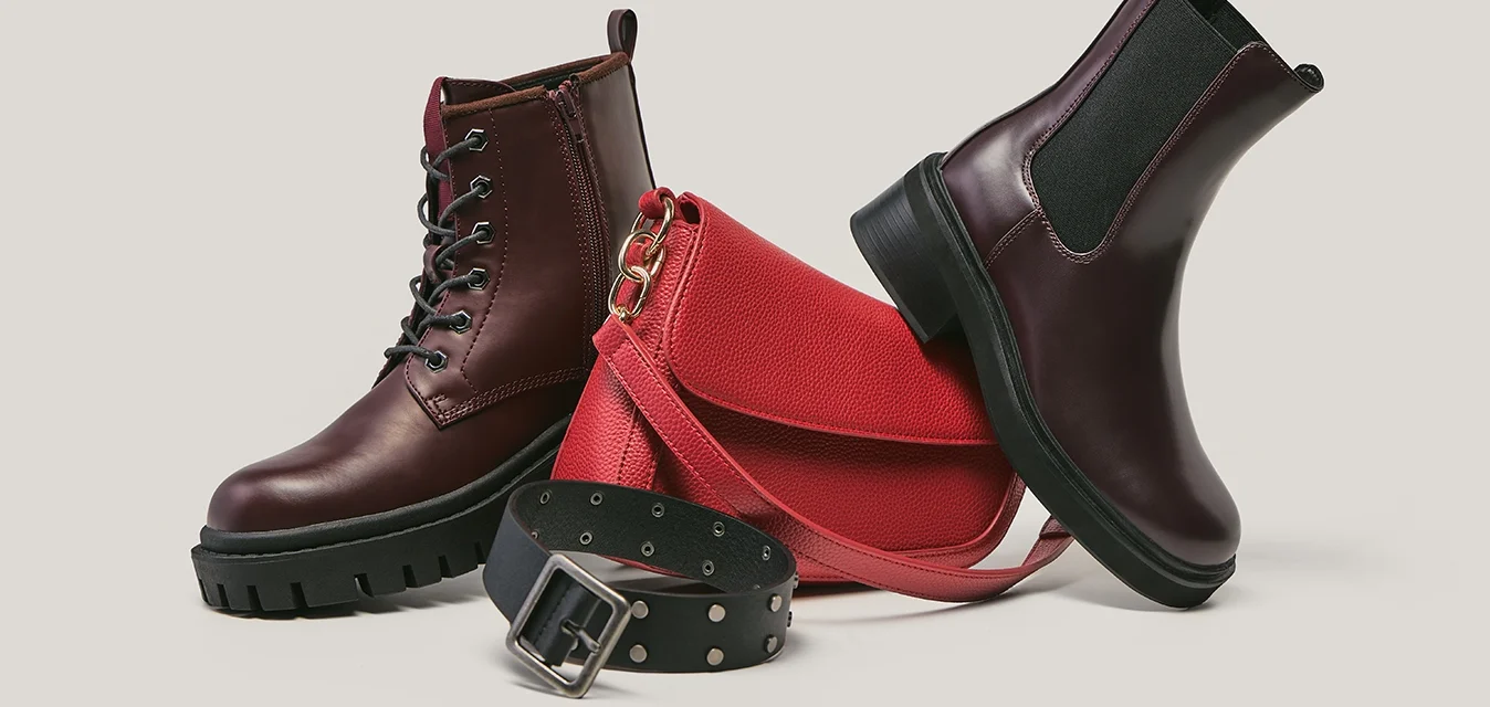 Stivale stringato in pelle marrone scuro e stivale Chelsea con suola nera, borsa a tracolla rossa con texture e cintura nera borchiata su sfondo chiaro.