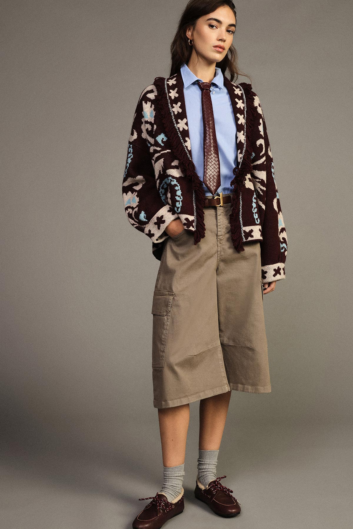 Young woman modelling patterned knitted cardigan over light-blue shirt and tie, khaki culottes and brown shoes