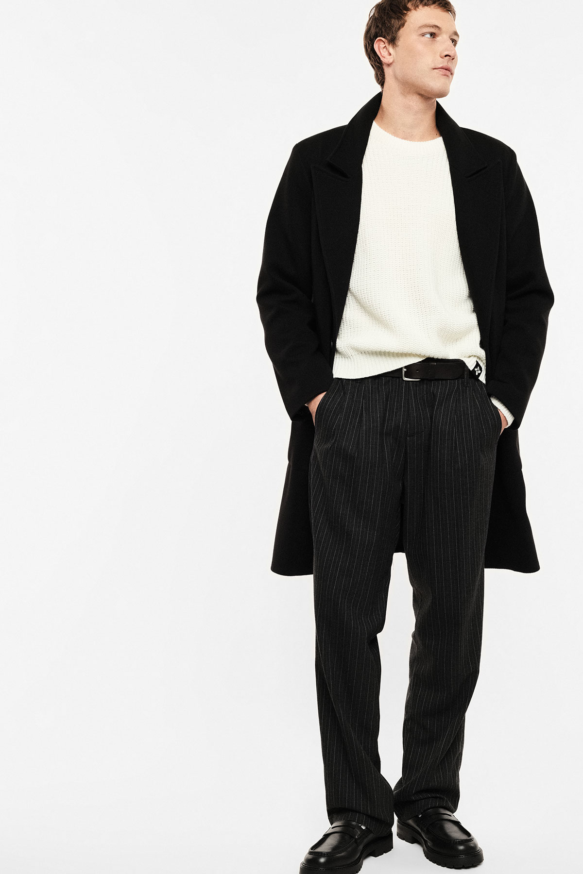 Male model wearing a black overcoat, cream knit sweater, pinstripe trousers and black leather loafers against a white background