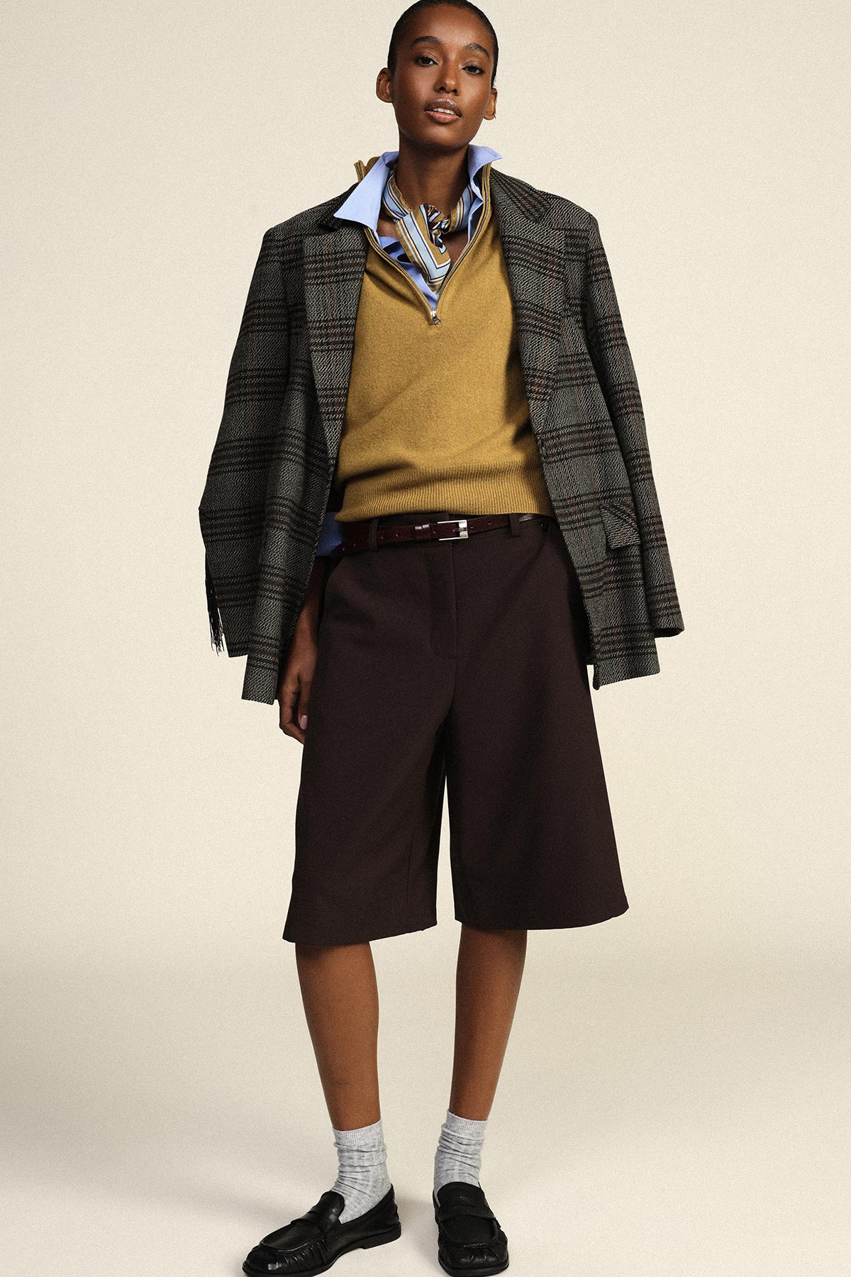 Model standing in a checked blazer draped over shoulders, mustard V‑neck jumper, striped shirt, dark long shorts, grey socks and black loafers
