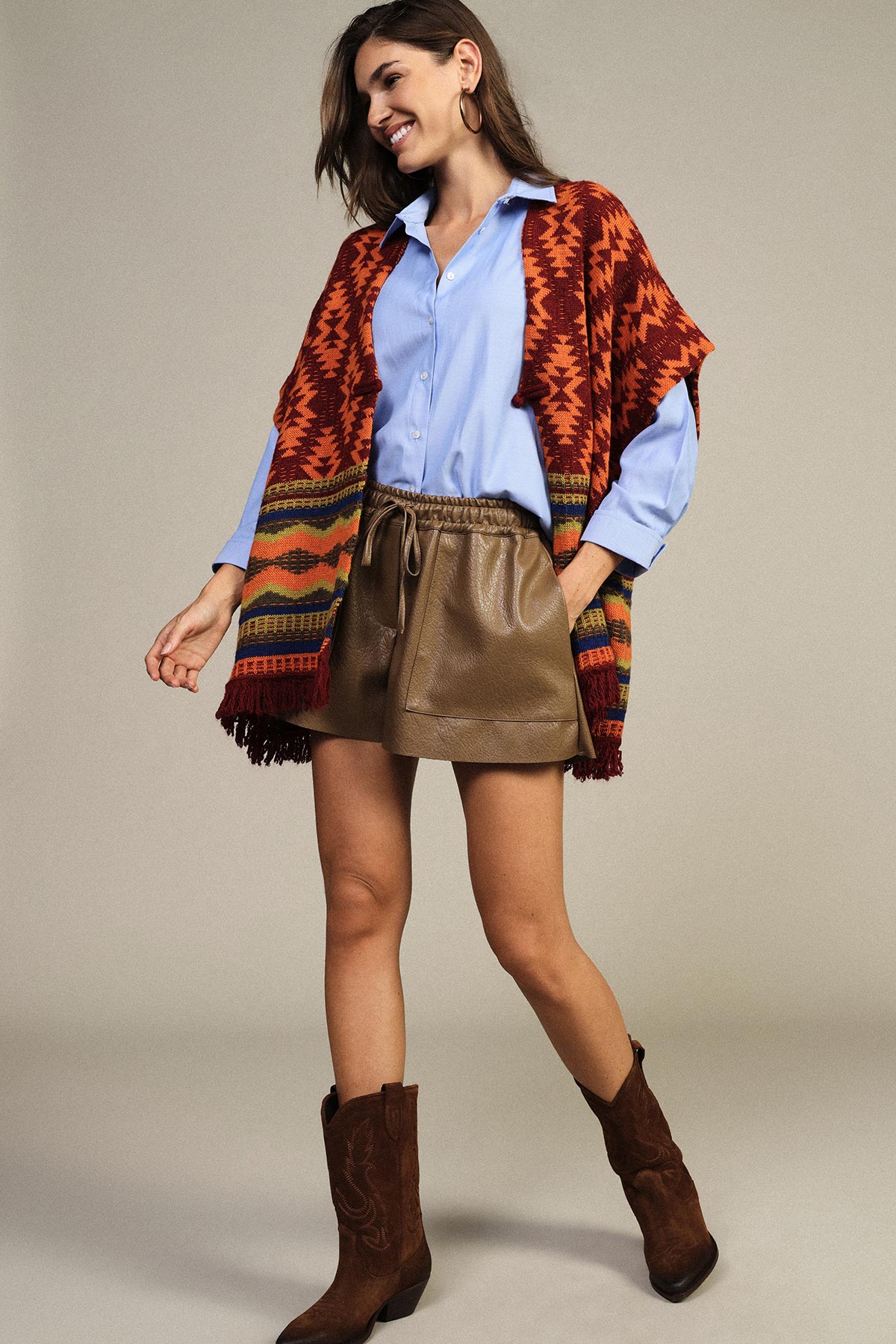 Woman wearing a rust patterned poncho, light blue shirt, brown leather shorts and suede cowboy boots, smiling with hands in pockets