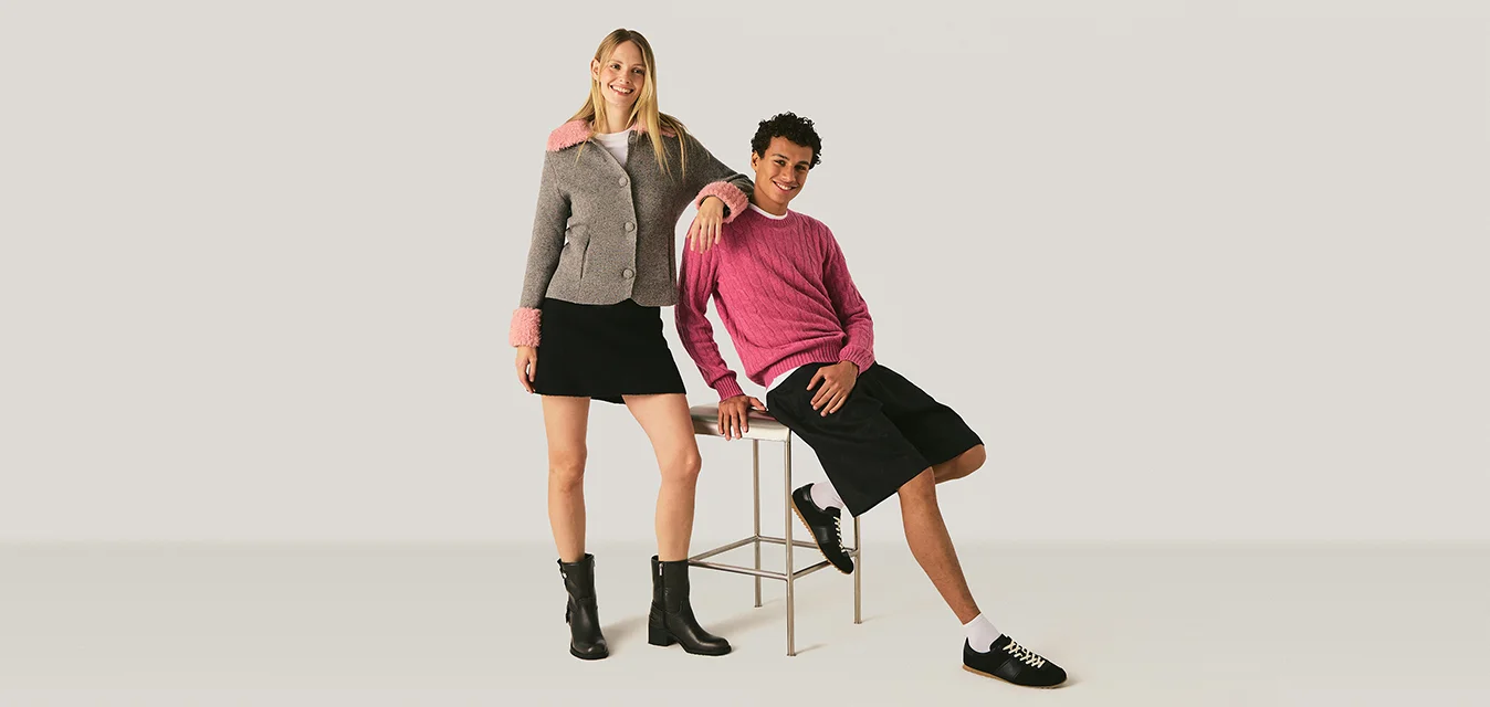 Giovane donna con giacca grigia e gonna nera in piedi accanto a un giovane con maglione rosa e pantaloncini neri seduto su uno sgabello su sfondo uniforme.