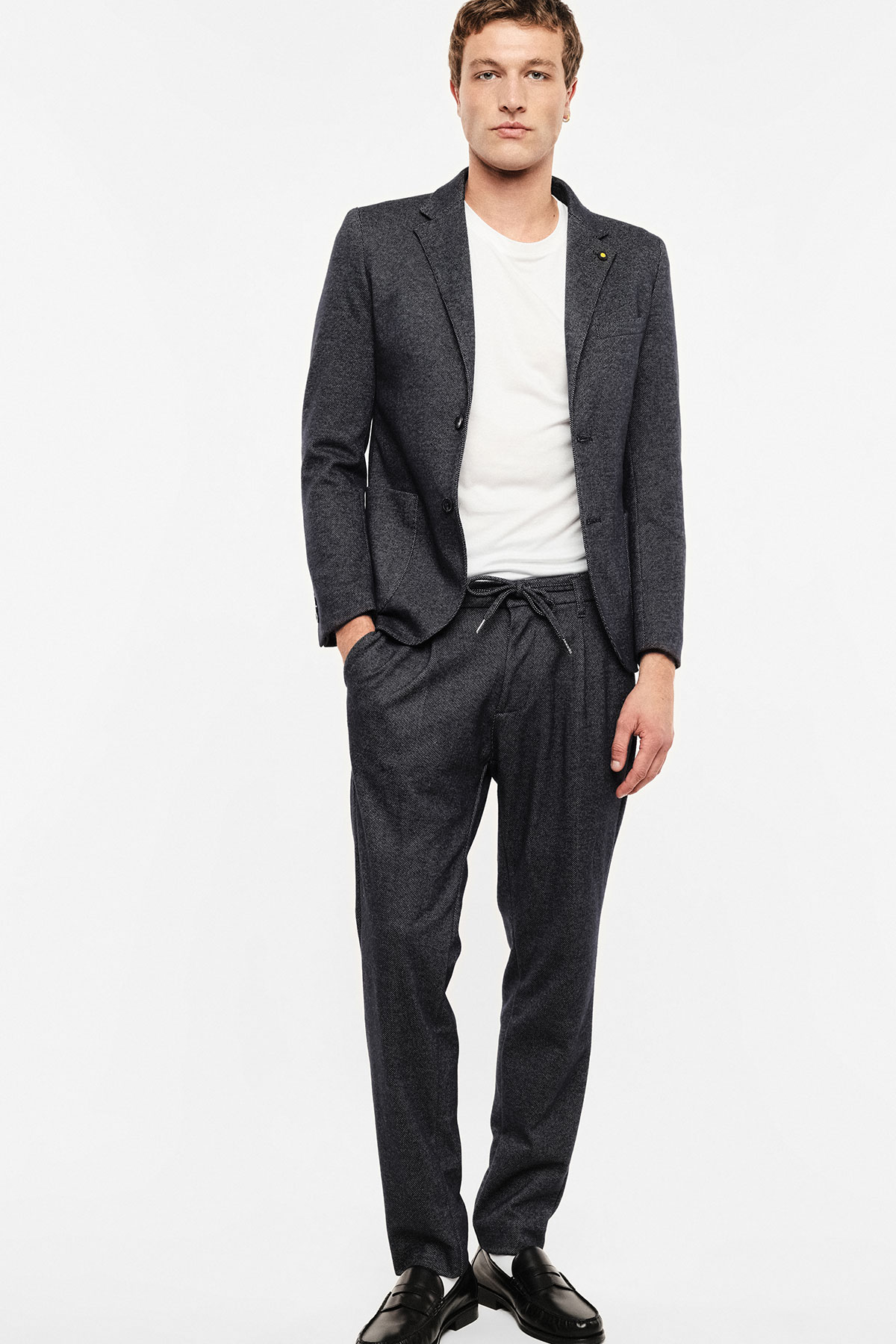 Young man wearing charcoal textured blazer and drawstring trousers with white T-shirt and black loafers, standing against a white background