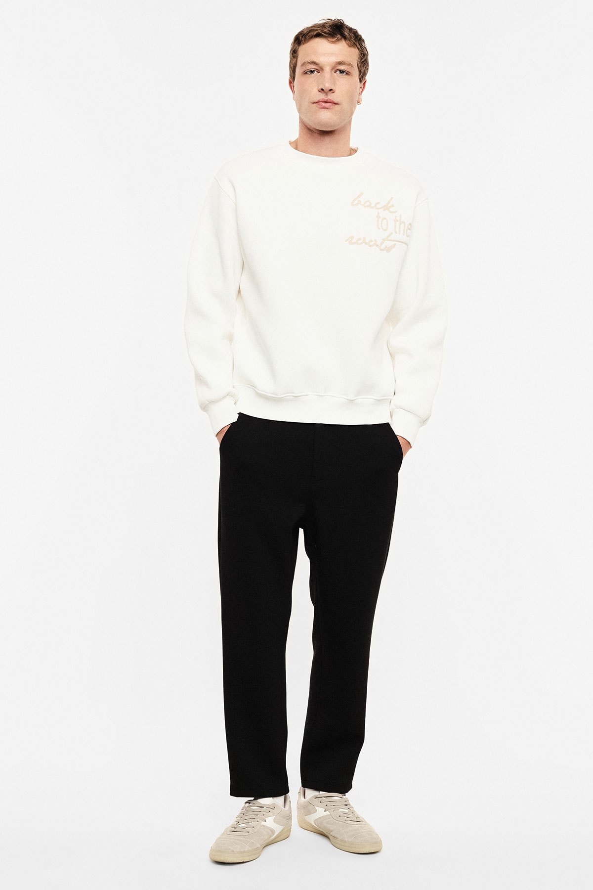 Man standing on white background wearing cream sweatshirt with script, black trousers and beige trainers, hands in pockets
