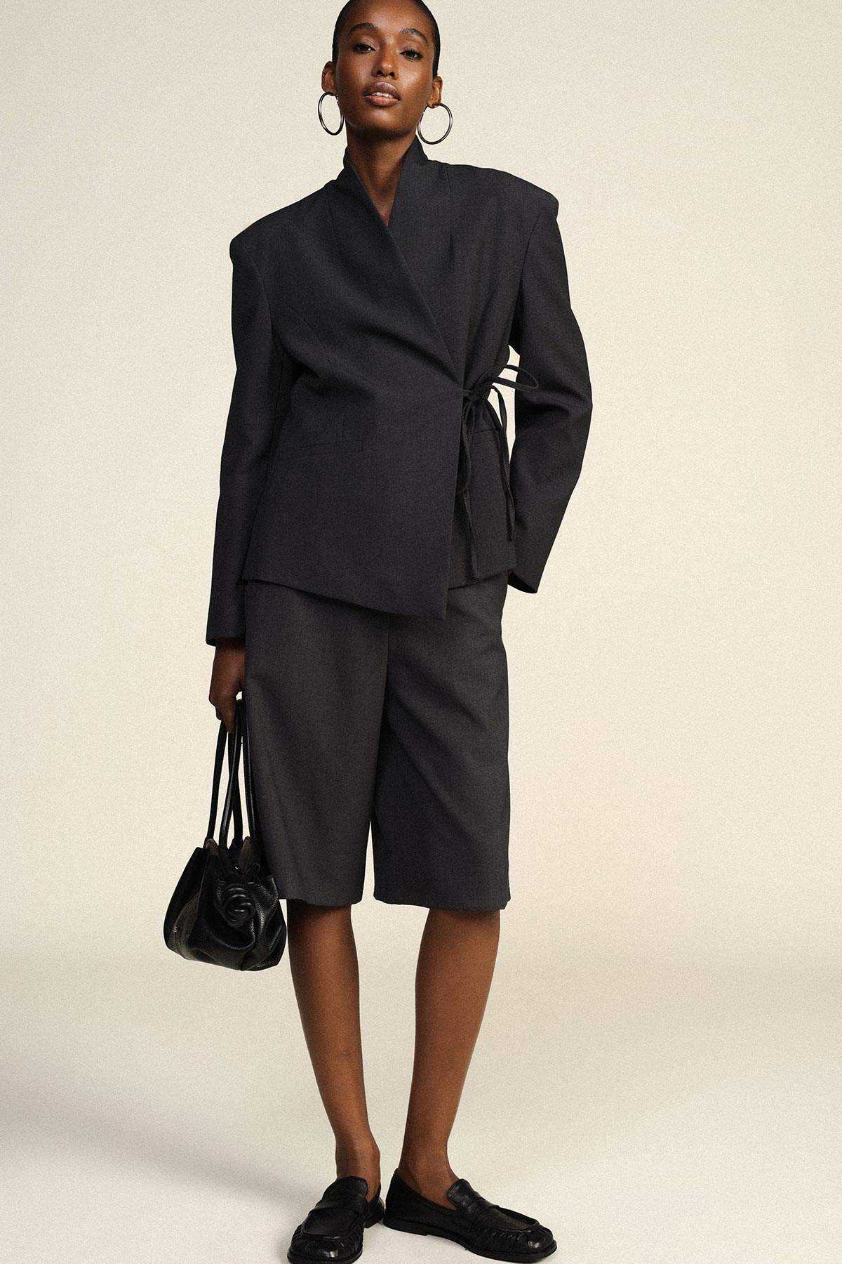 Black wrap blazer with side tie, matching knee-length shorts, black loafers and handbag on model with hoop earrings