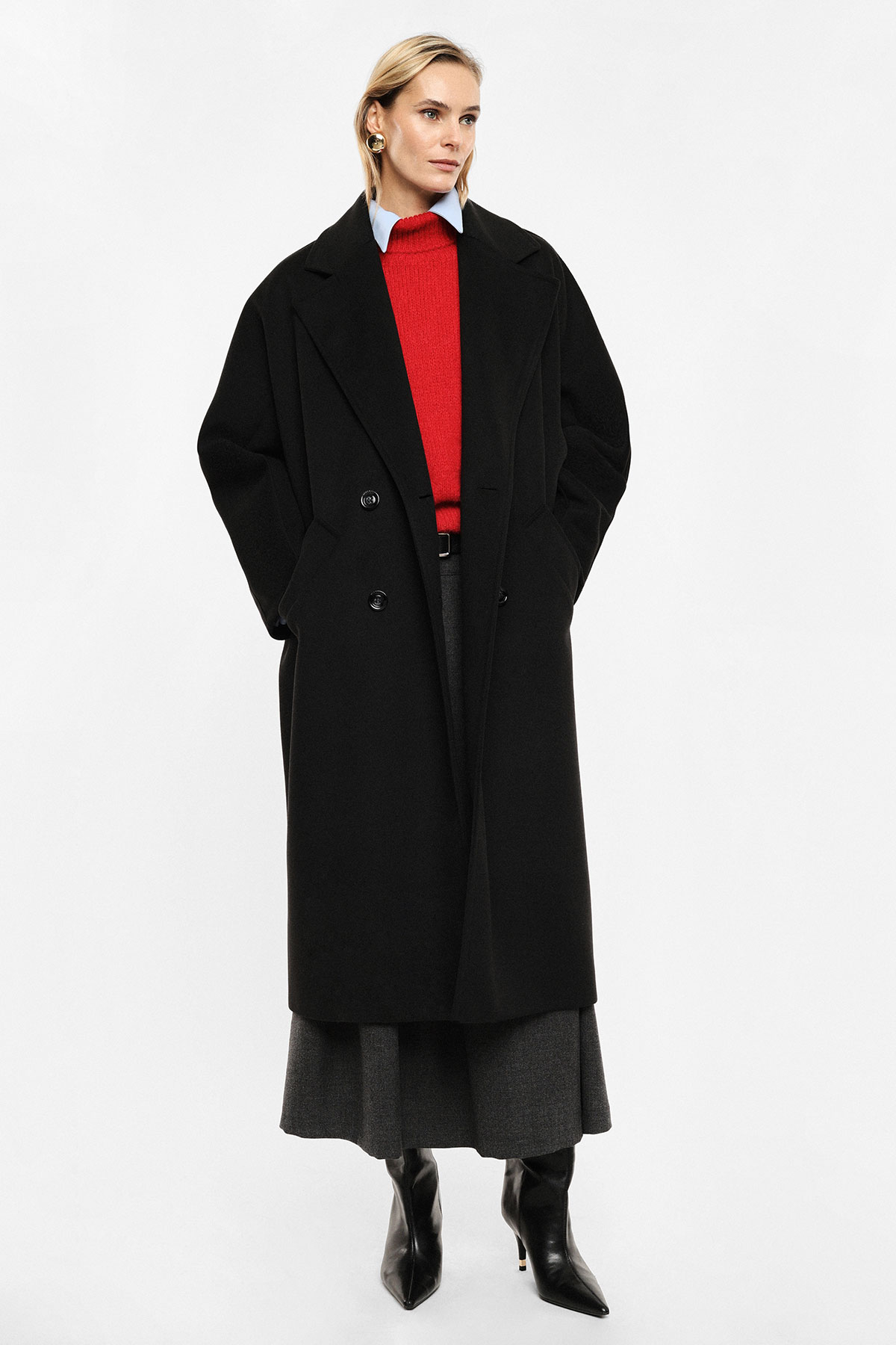 Woman wearing long black overcoat over a red sweater, grey skirt and black heeled boots on white background
