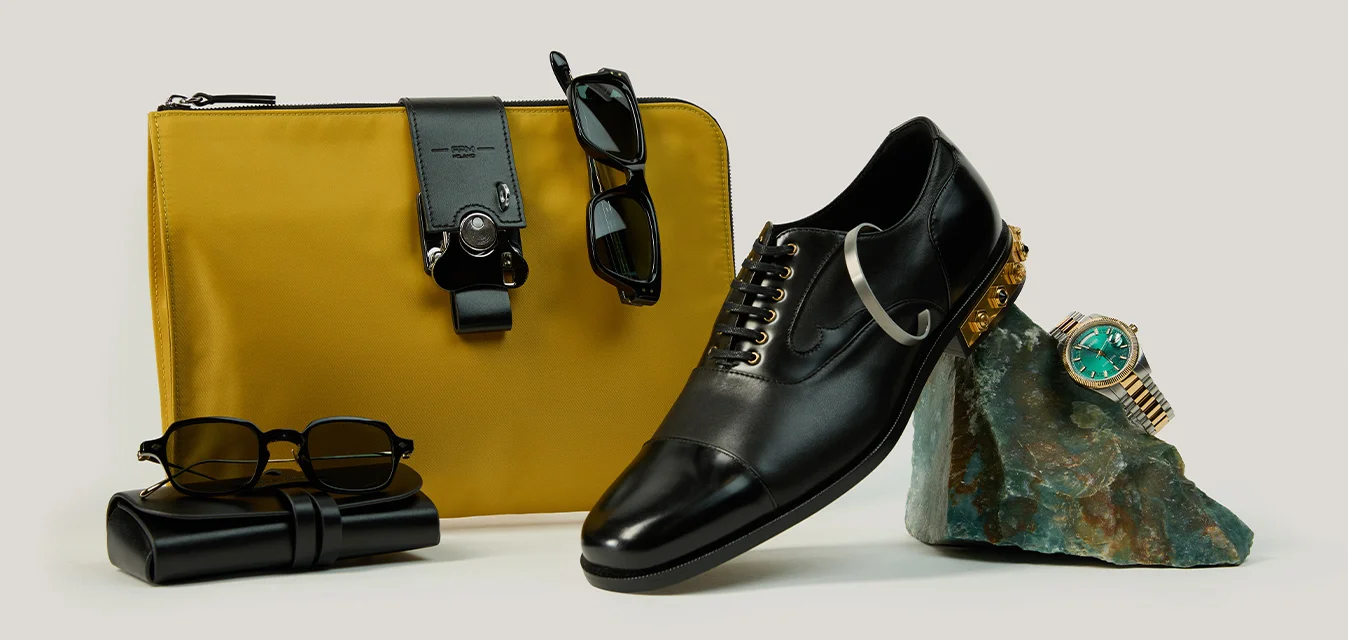 Scarpa elegante in pelle nera appoggiata su una roccia verde con un orologio da polso, borsetta gialla con occhiali da sole e astuccio nero piegato per occhiali su sfondo bianco.