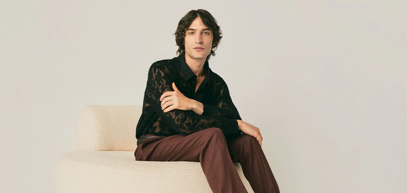 Giovane seduto su un pouf tondo beige, indossando una camicia di pizzo nera, pantaloni bordeaux, calze decorate e scarpe di pelle marroni in una stanza minimalista.