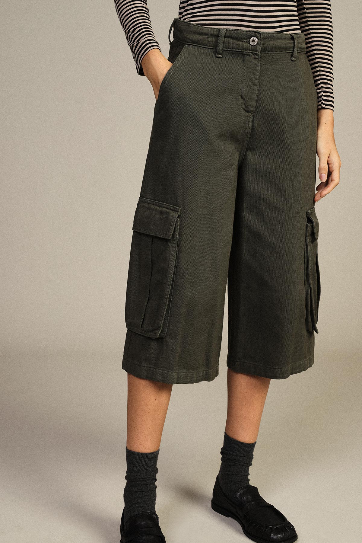 Waist-down view of a model wearing olive-green cargo culotte shorts with side flap pockets, striped long-sleeve top, dark socks and black loafers