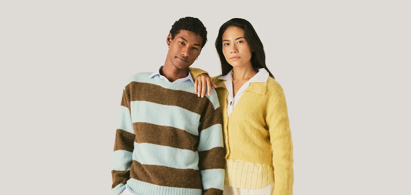 Due adulti sono insieme davanti a uno sfondo semplice; uno indossa un maglione a righe blu e marrone, l'altro un cardigan giallo sopra una camicia bianca.