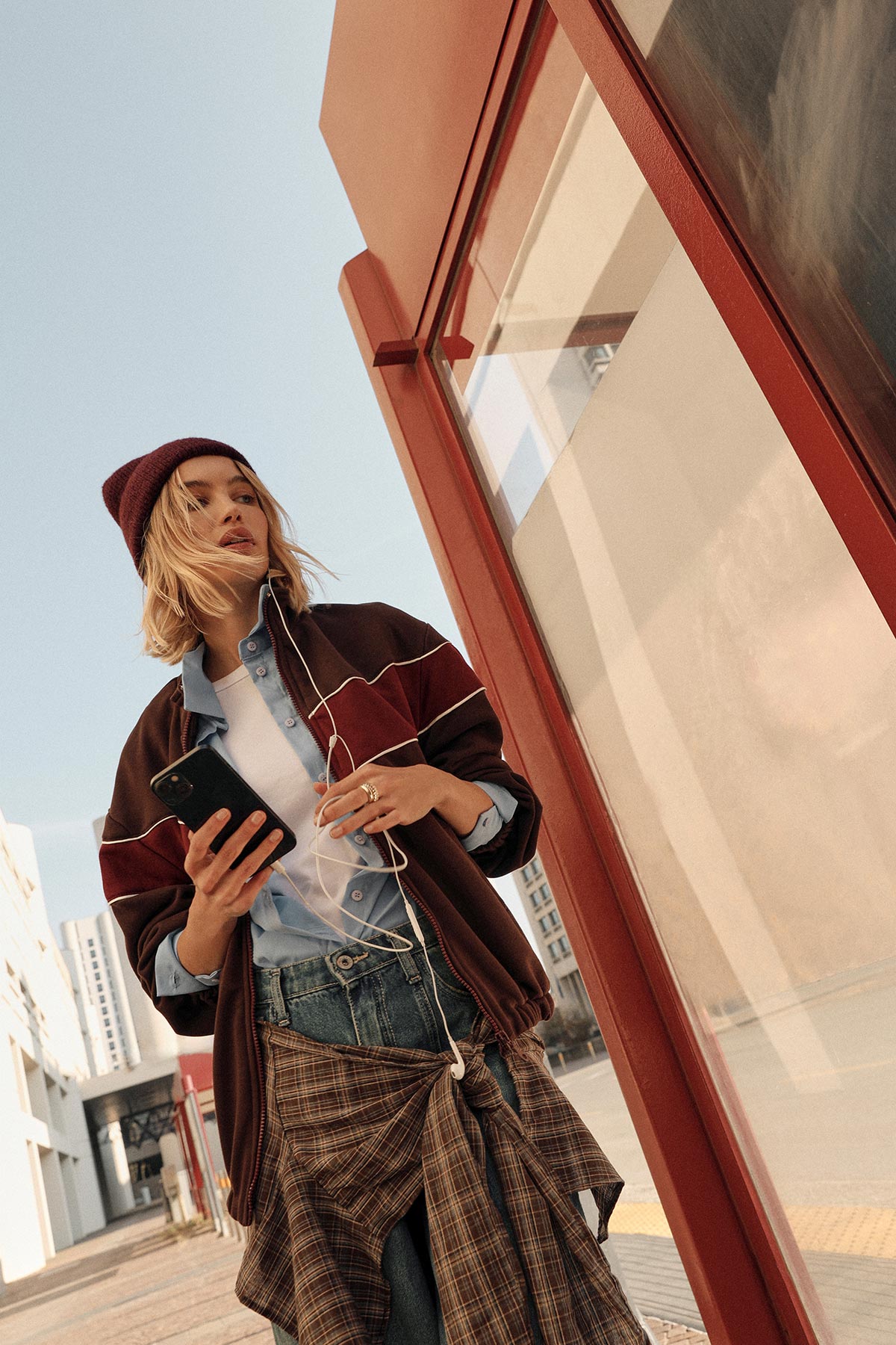 Young woman in a beanie and layered casual clothes walking in a city street, holding a smartphone with earphones